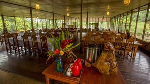 A spacious dining area with wooden tables and chairs, large windows, and a decorative centerpiece on the table. - Pokakempi Tours (Tambopata)