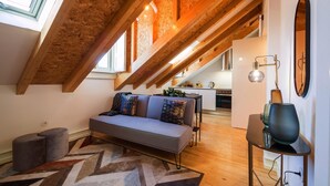 Un salon de style industriel, avec des poutres en bois apparentes, un canapé gris agrémenté de coussins à motifs, un tapis géométrique et une lampe moderne posée sur une table d’appoint.