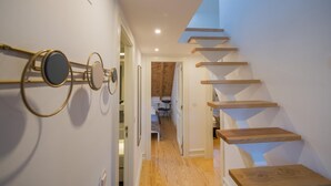 Un escalier moderne avec des marches en bois et une structure en verre. Un couloir avec un plancher en bois et un mur blanc orné d’un miroir rond.