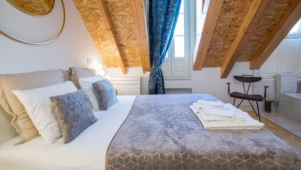 Une chambre à coucher avec un lit, des oreillers, une fenêtre avec des rideaux, une chaise et une petite table.