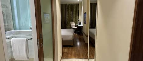 A hotel corridor with a bathroom on the left, a bedroom with a bed and a desk, and a mirror reflecting the room.