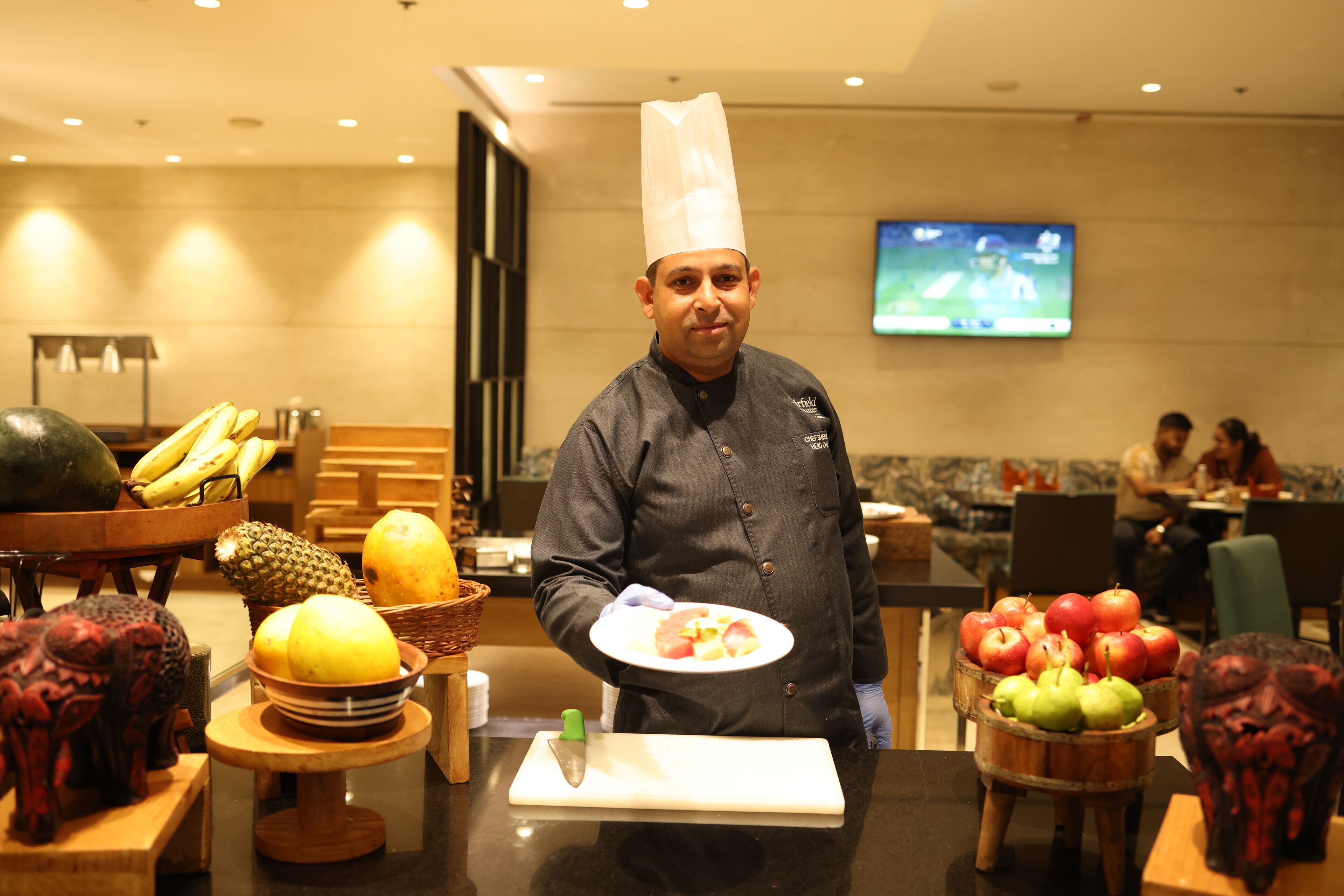 A chef in a uniform holding a plate of food, with fruit baskets and a cutting board on the counter.