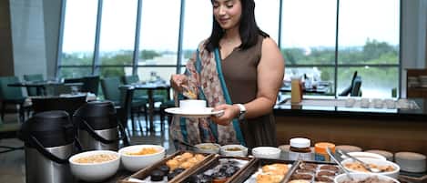 Montaje tipo buffet con diversos alimentos y una estación de café en un comedor moderno con grandes ventanales.