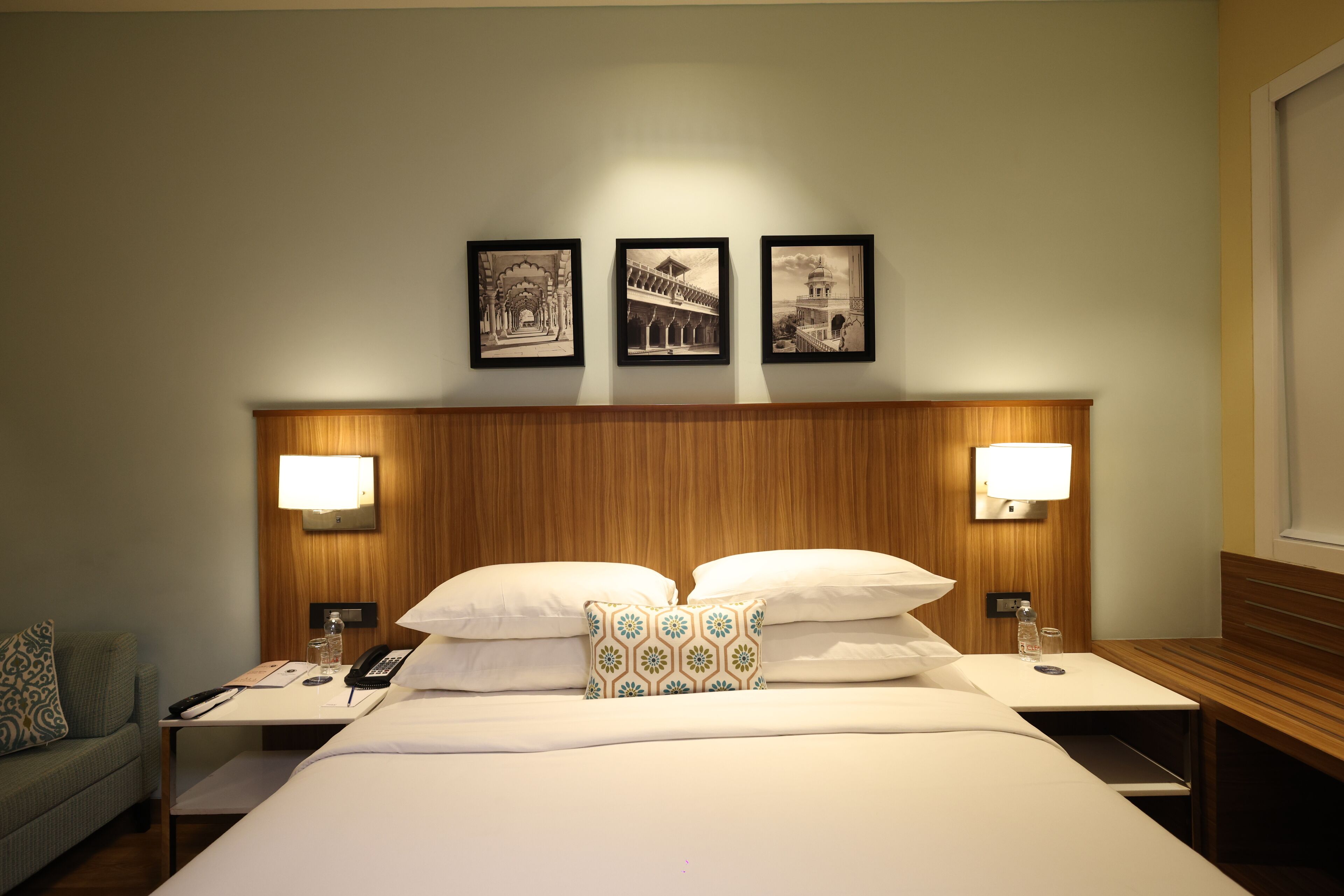 A hotel room with a large bed, two bedside tables, a nightstand, a lamp, a telephone, a water bottle, and three framed pictures on the wall.