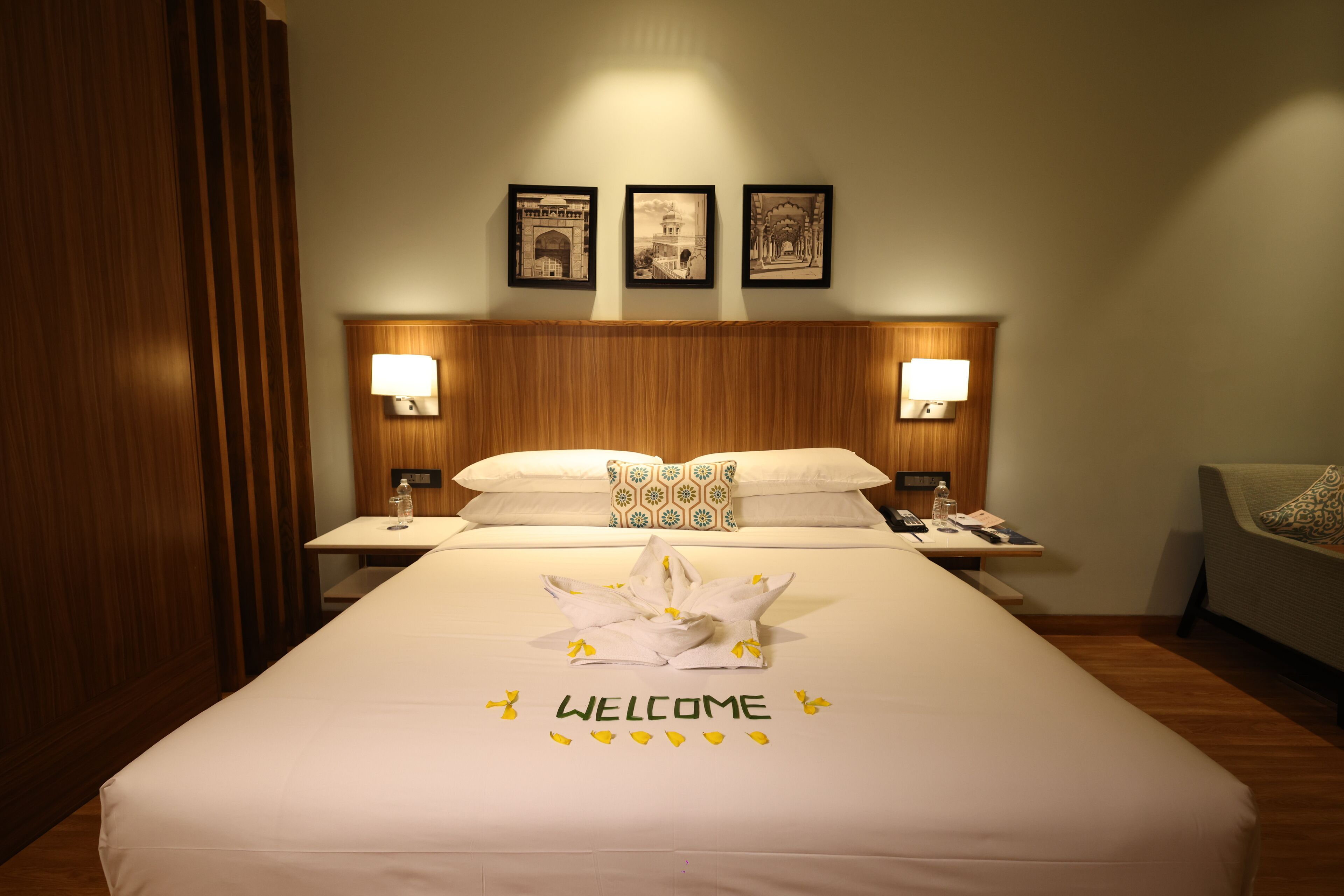 A hotel room with a neatly made bed, a bedside table with a lamp, a small table with a bottle, and a wall-mounted picture frame.