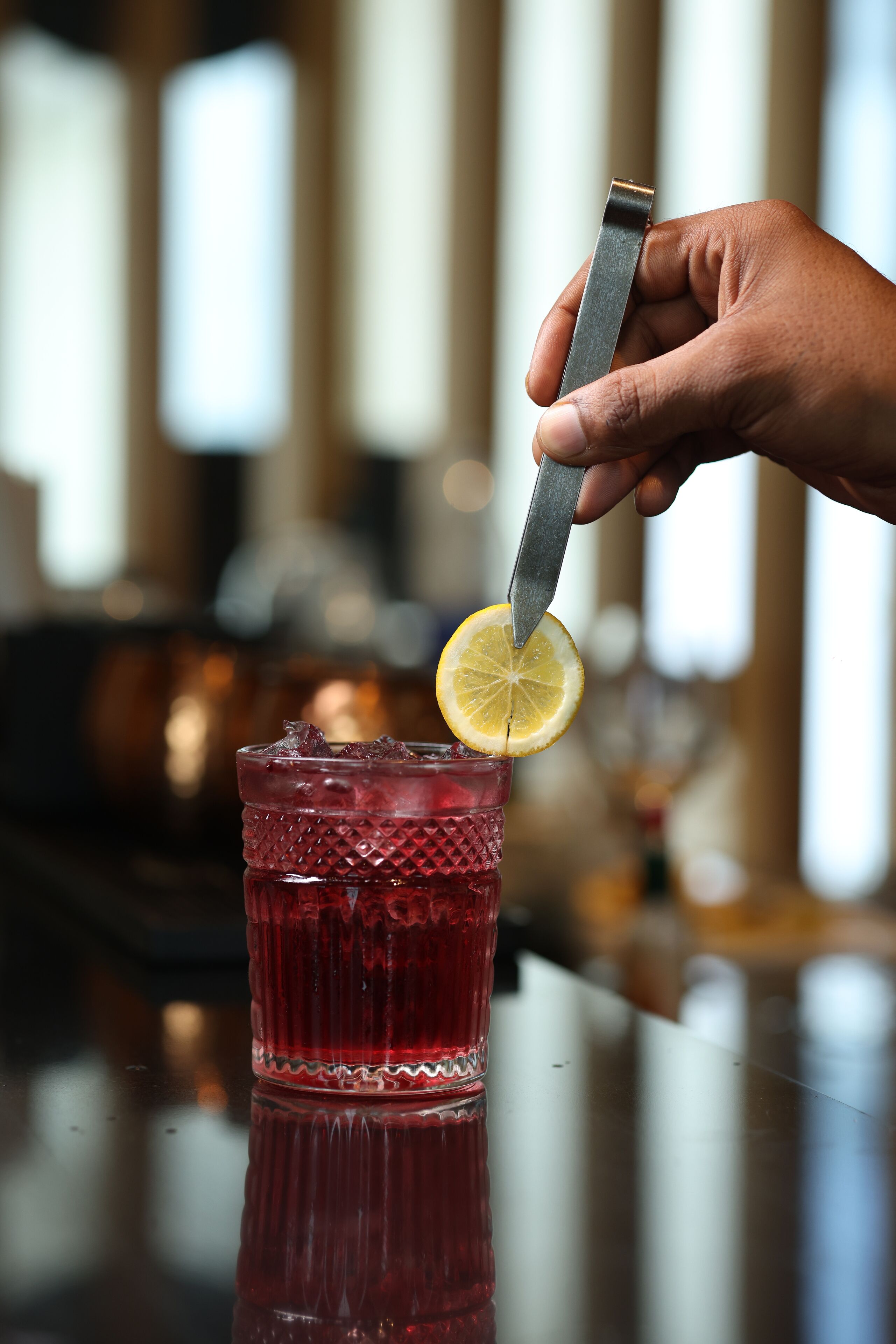 A hand holding a knife, cutting a lemon slice into a glass of red beverage.