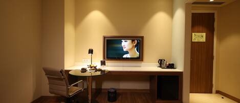 A hotel room with a wooden floor, a desk with a chair, a television mounted on the wall, and a small trash bin.