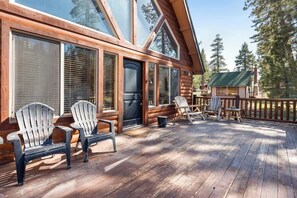 A wooden deck with Adirondack chairs, large windows, and a small building in the background.