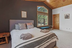 A bedroom with a large bed, a wooden bench, a window with a view of trees, and a door. - Wolf Den Cabin • Spa • Walk to Lake (Big Bear Lake)