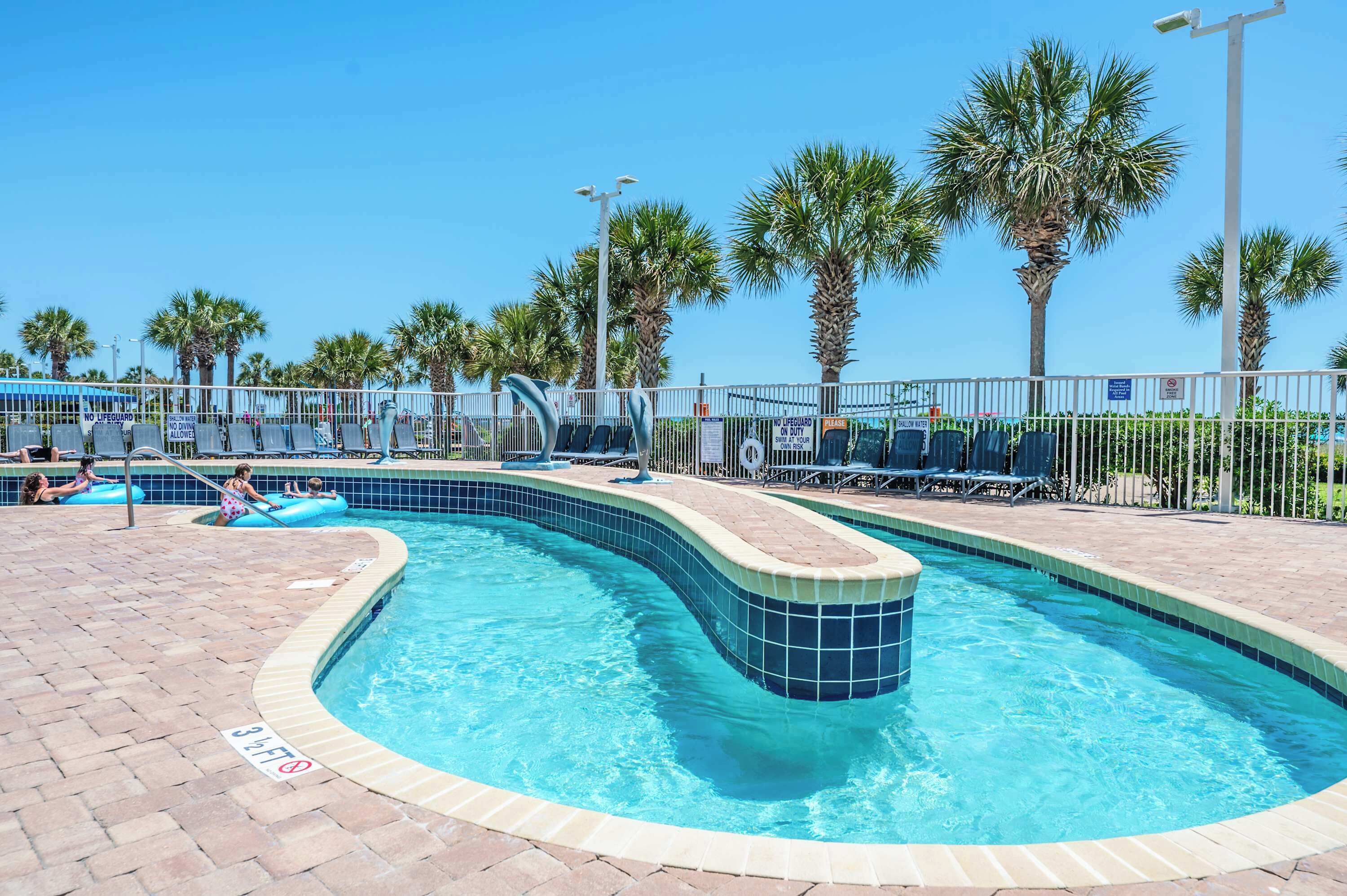 A swimming pool with a tiled edge, palm trees, and a fenced area with lounge chairs.