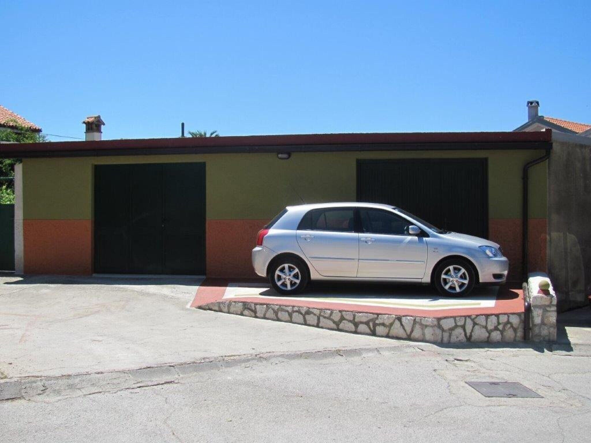 Une voiture argentée est garée devant un immeuble dont la porte du garage est visible, ainsi qu’une petite construction en pierre située à l’avant.