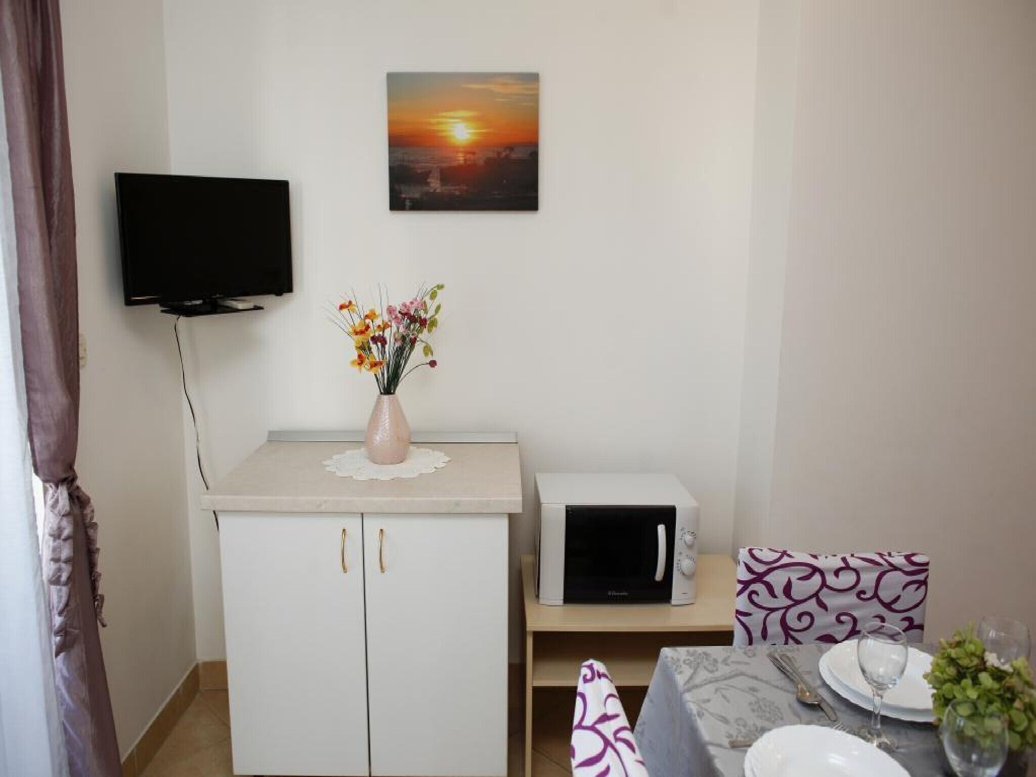 A small kitchenette with a microwave, a sink, and a cabinet. There is a flat-screen TV mounted on the wall above the counter.