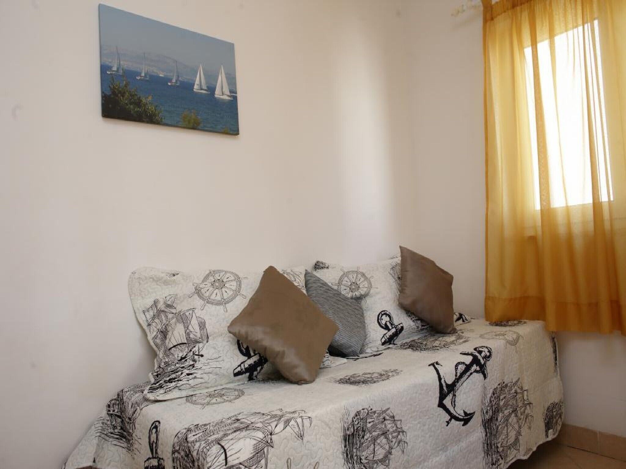 Habitación con sofá cama, ropa de cama con motivos náuticos, un cuadro de veleros en la pared y una ventana con cortinas traslúcidas.