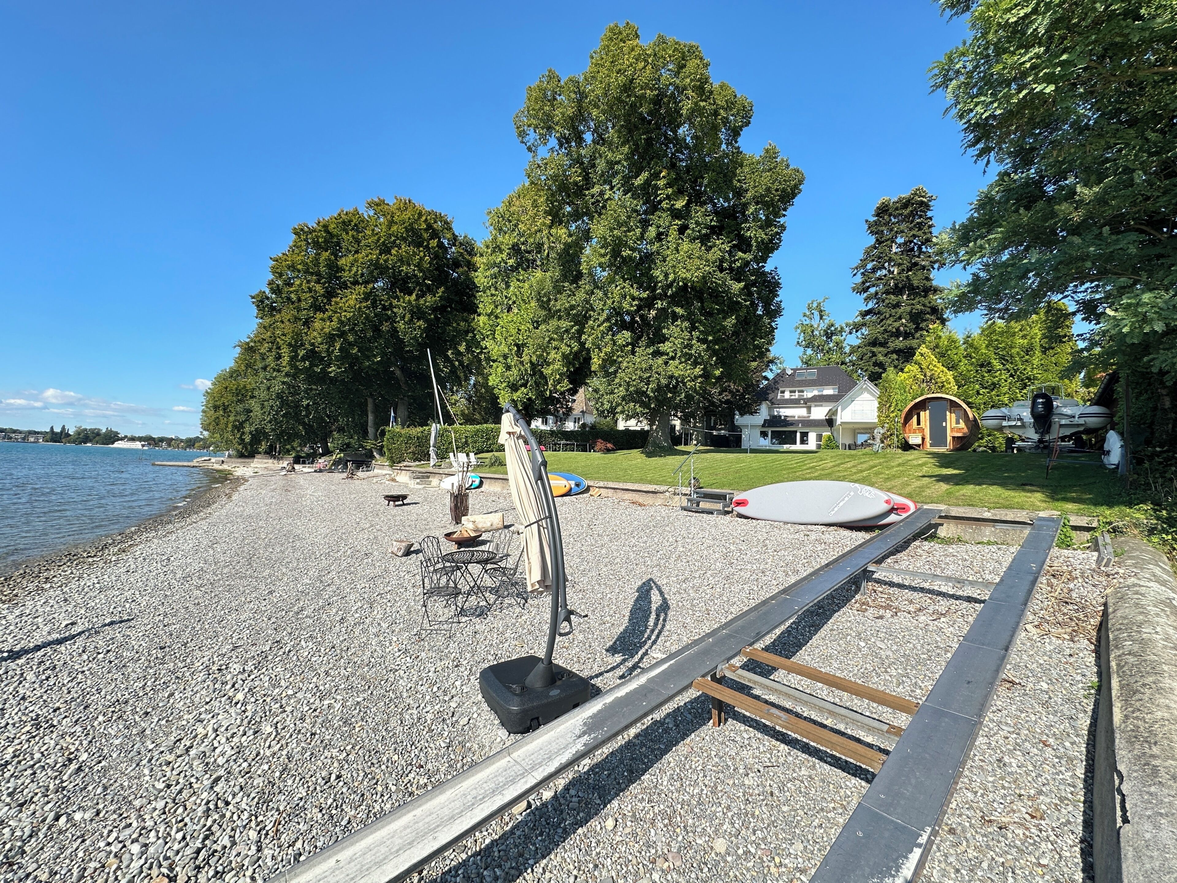 Een kiezelstrand met een jetski, een ligstoel en een klein bootje afgemeerd nabij een huis met een gazon.