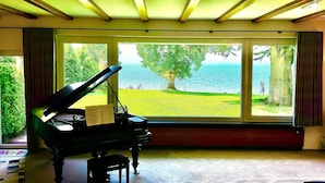 A grand piano in a room with large windows overlooking a grassy area and water. - Lakefront vacation home with private beach & sauna – Villa Noho (Nonnenhorn)