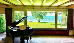 A grand piano in a room with large windows overlooking a grassy area and water.