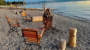 A pebbled beach with wooden benches and a fire pit. - Lakefront vacation home with private beach & sauna – Villa Noho (Nonnenhorn)