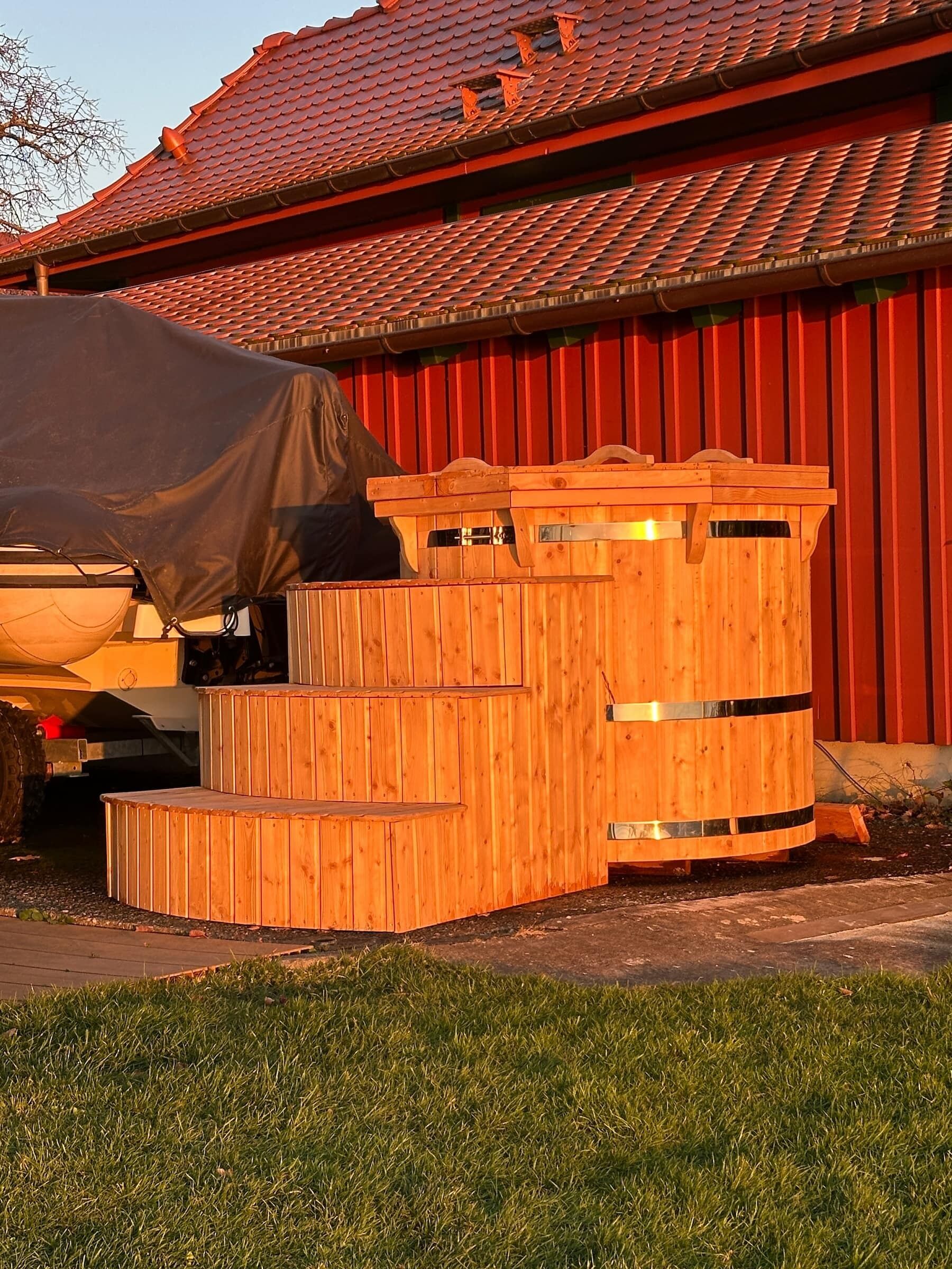 A wooden outdoor structure with steps and a barrel, likely a barbecue area, next to a red building with a tiled roof.
