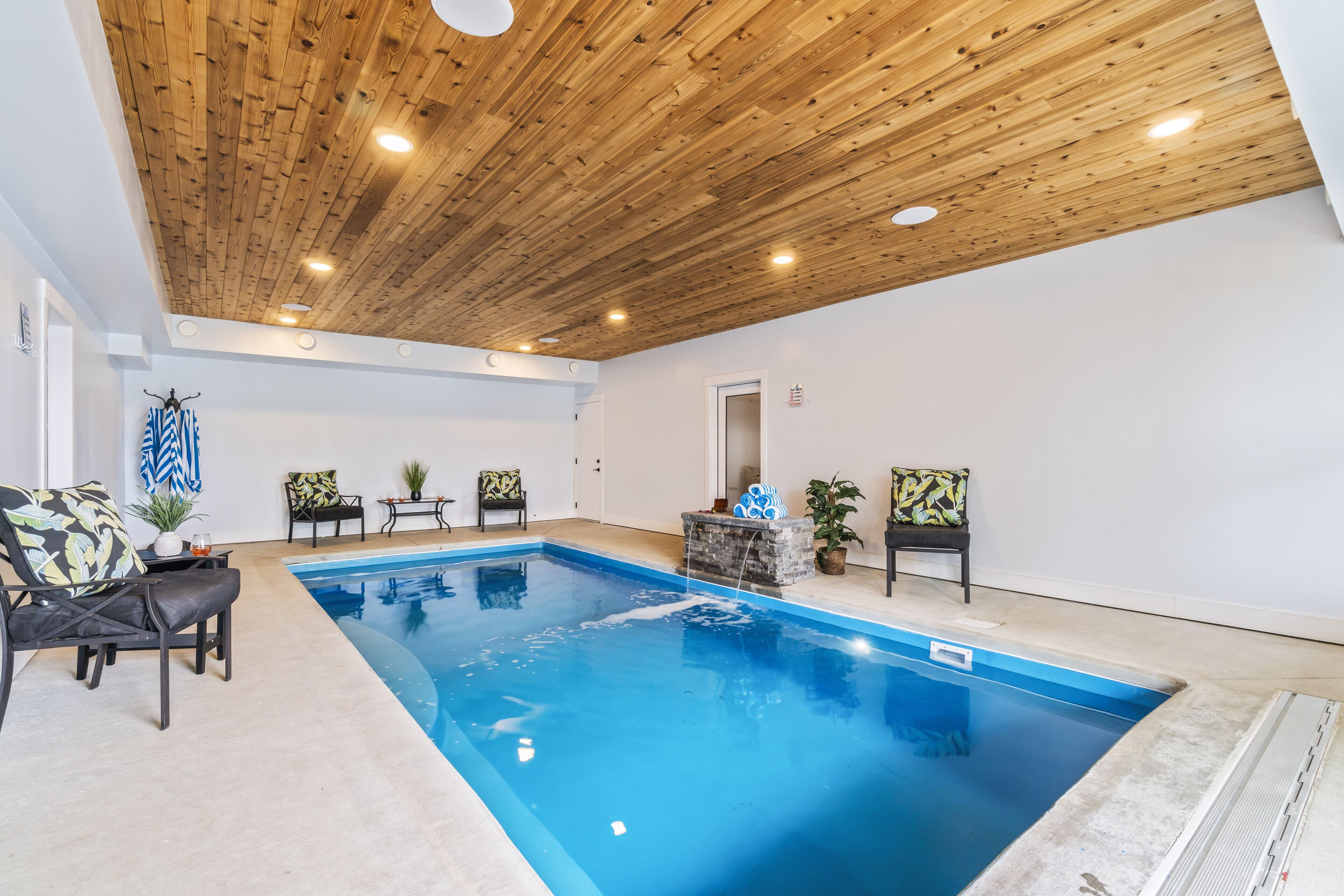 An indoor swimming pool with wooden ceiling beams, lounge chairs, and potted plants.