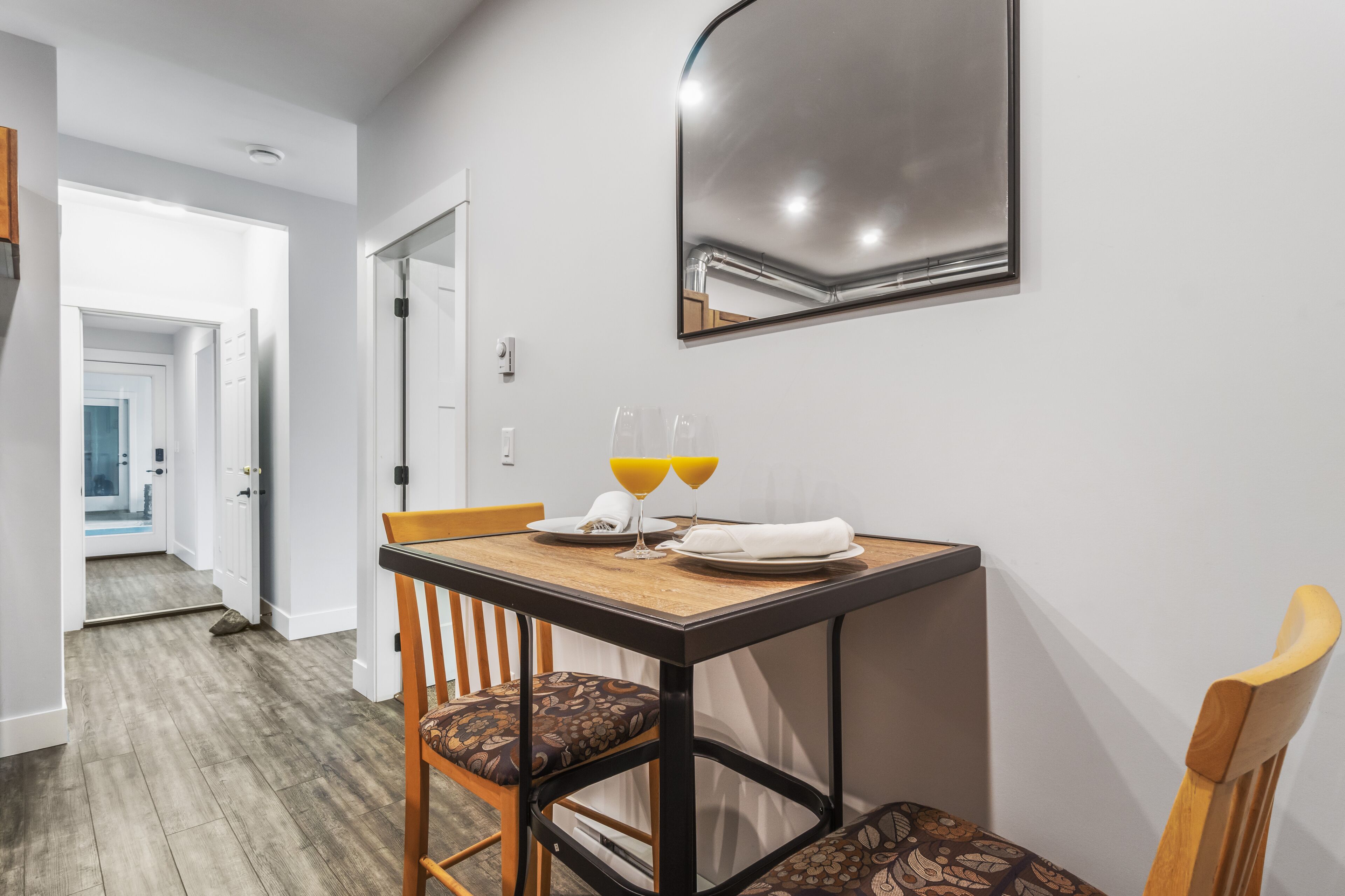 Un área de comedor con una mesa lista para dos personas, un espejo sobre la mesa y una puerta que da a otra habitación.