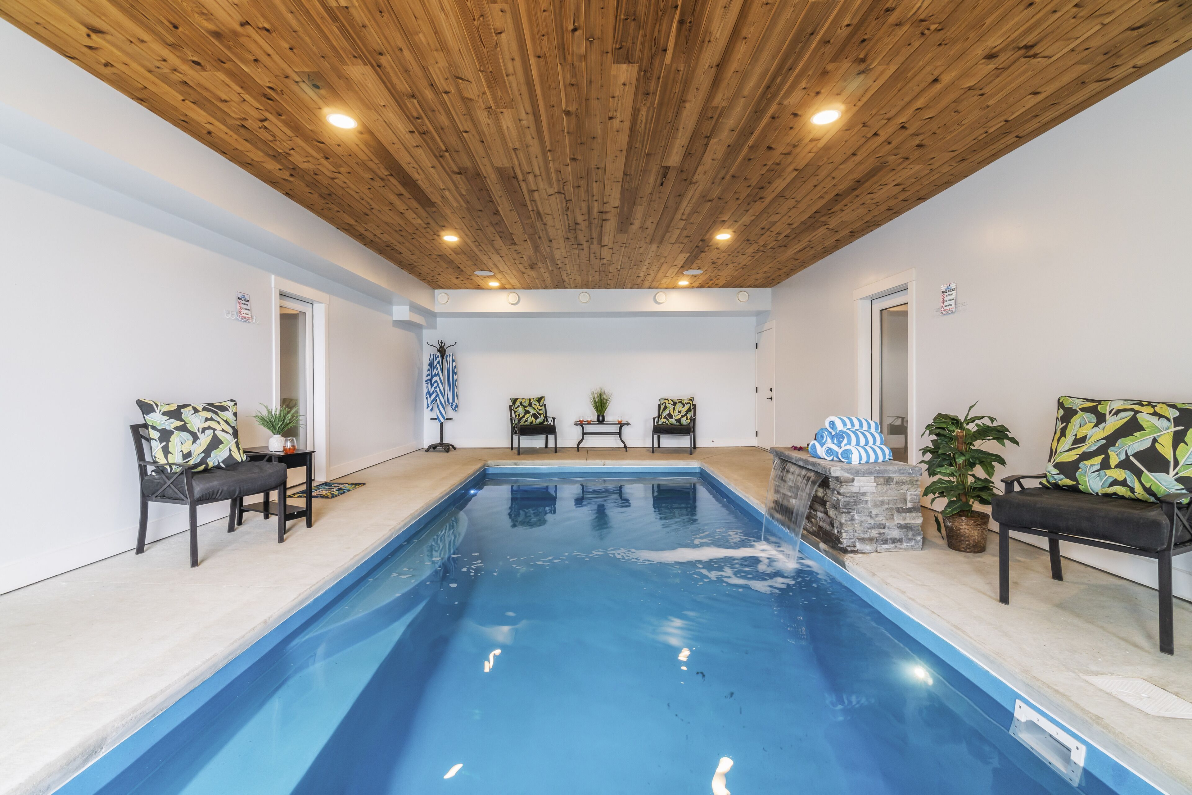 Une piscine intérieure avec un plafond en bois, deux fauteuils avec des coussins et une petite table sur laquelle est posée une plante.