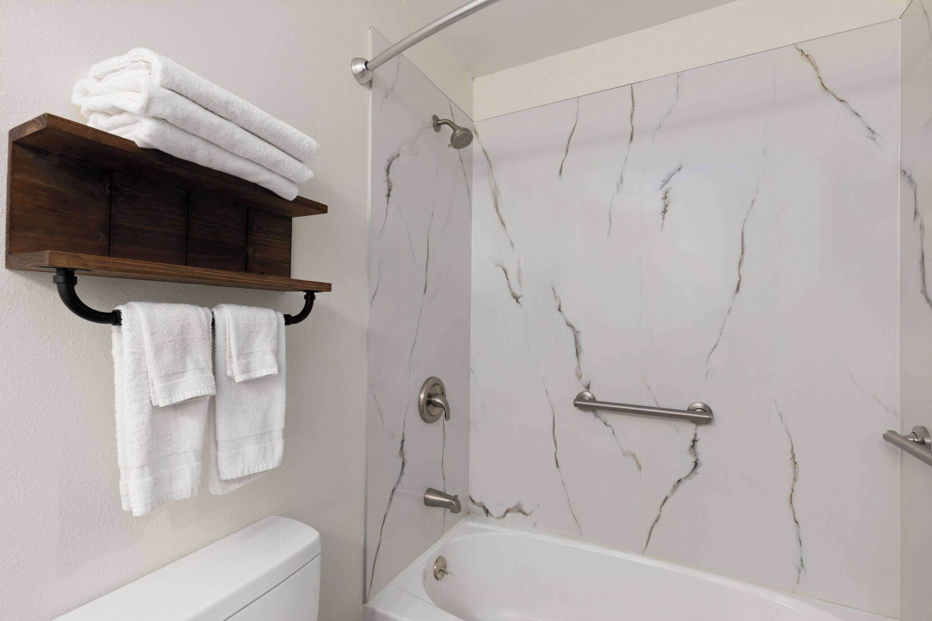 A bathroom with a bathtub, marble walls, a wooden shelf with towels, and a shower area.