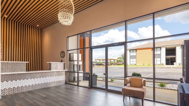 A reception area with a wooden reception desk, a chair, and large windows offering a view of the parking lot and buildings outside. - Ramada by Wyndham Studio Suites Dothan (Dothan)
