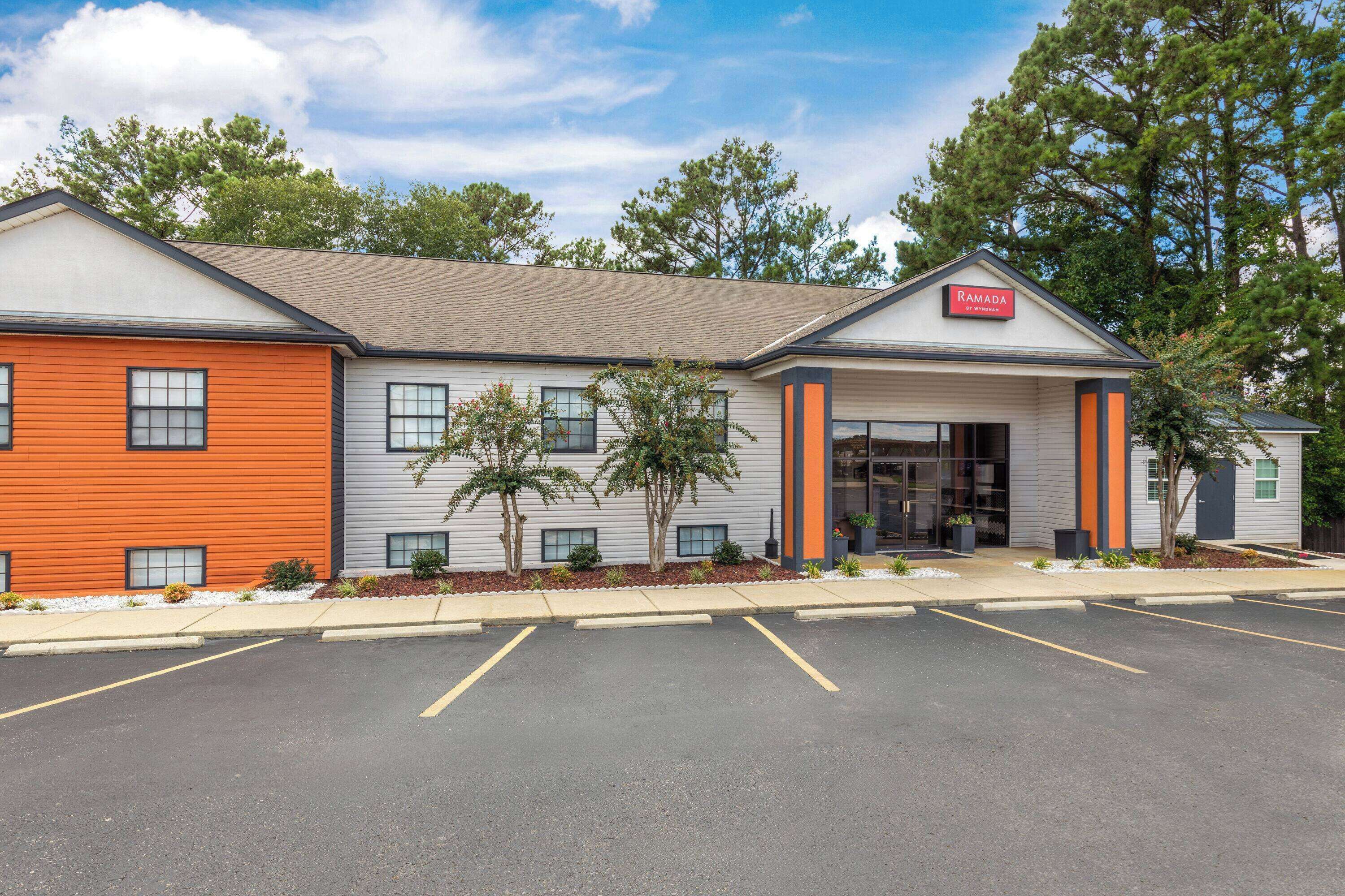 A Ramada hotel with a modern design, featuring a mix of orange and grey exterior walls, a covered entrance, and a parking lot.