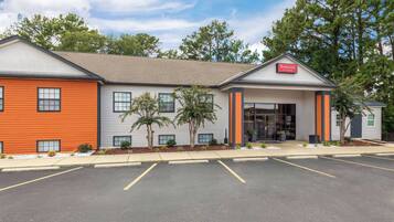 A Ramada hotel with a modern design, featuring a mix of orange and grey exterior walls, a covered entrance, and a parking lot.