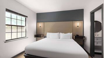 Una habitación de hotel moderna con una cama grande, dos mesitas de noche, un tocador, una ventana y un baño con lavabo y espejo.