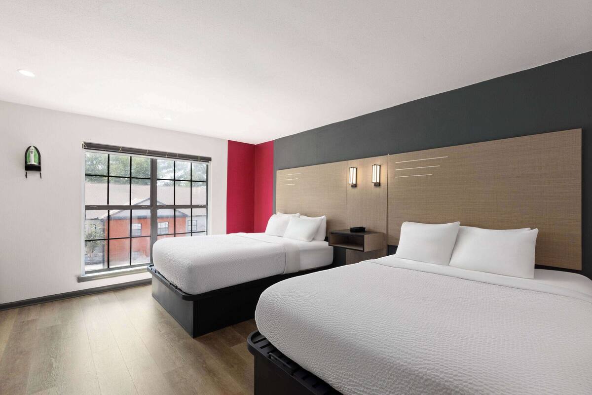 A hotel room with two beds, a large window, a red accent wall, and a modern design.