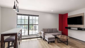 A modern living room with a sofa, ottoman, coffee table, and a dining area with a table and chairs. - Ramada by Wyndham Studio Suites Dothan (Dothan)