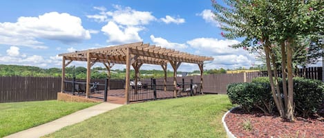 Een houten pergola met een patio tafel en stoelen, omringd door een gazon en een decoratieve rotstuin.