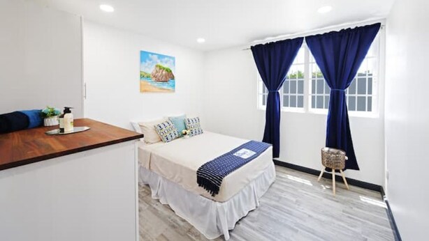 Une chambre à coucher avec un lit, un bureau en bois, une petite table et une fenêtre avec des rideaux.