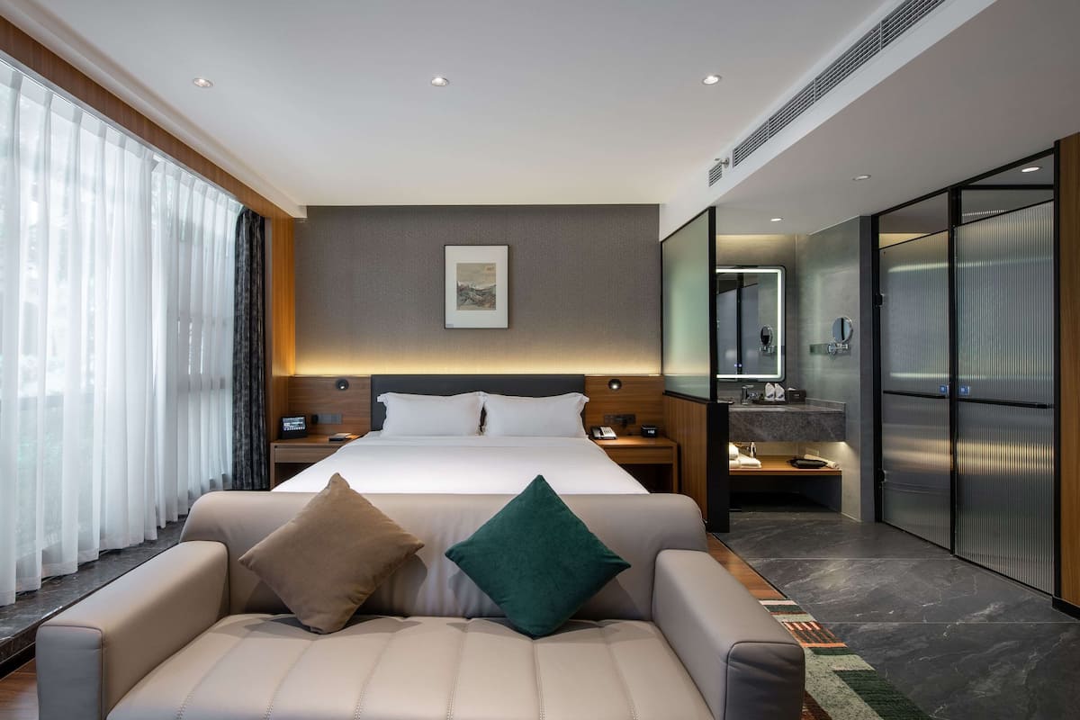 A modern hotel room with a large bed, a sofa, a bedside table, a nightstand, a mirror, and a wardrobe.