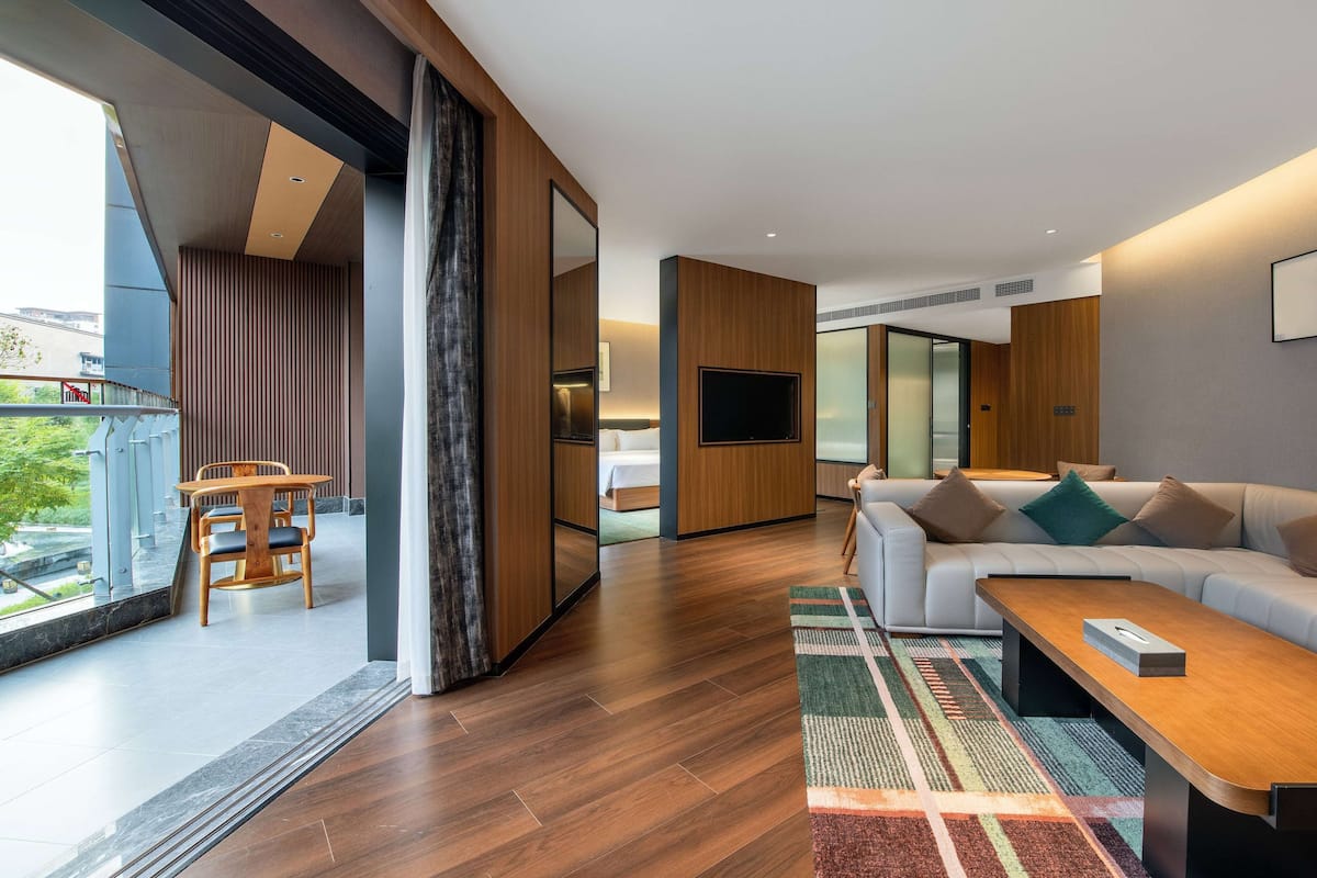A modern living room with a sofa, coffee table, and a TV. There is a dining area with a table and chairs. The room has wooden flooring and large windows.