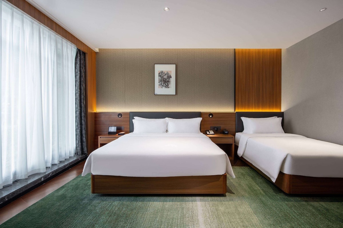 A modern hotel room with a large bed, two bedside tables, a nightstand, and a large window with curtains.