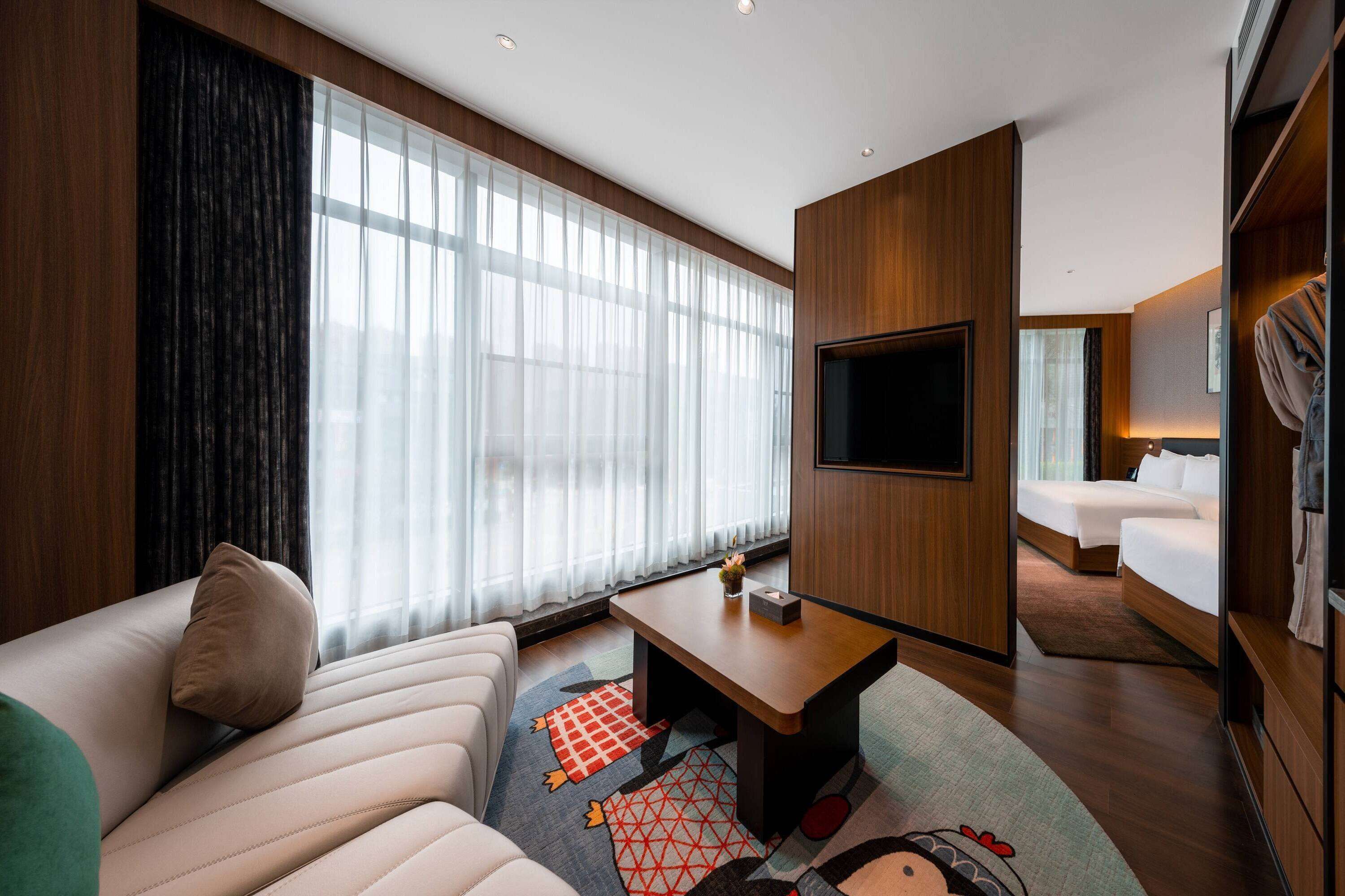 A modern hotel room with a large bed, a sofa, a TV, and a window with sheer curtains.