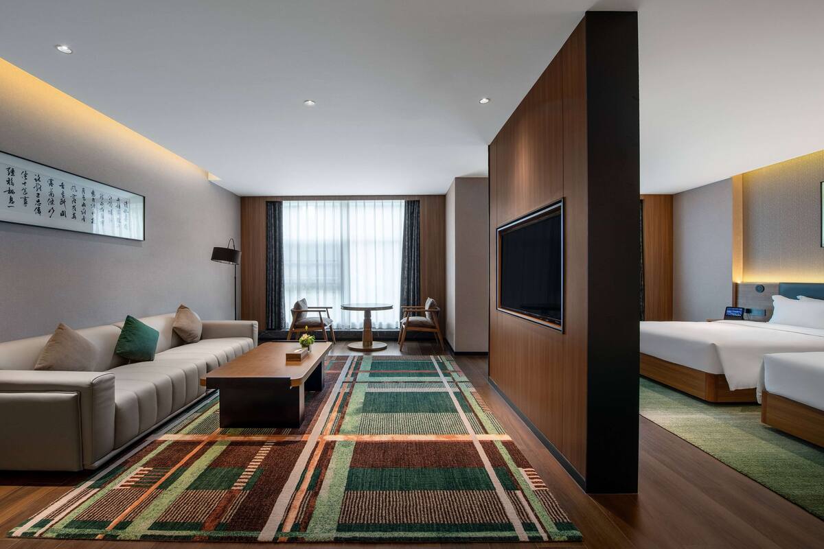 A modern hotel room with a sofa, a coffee table, a TV, a bed, and a large window.