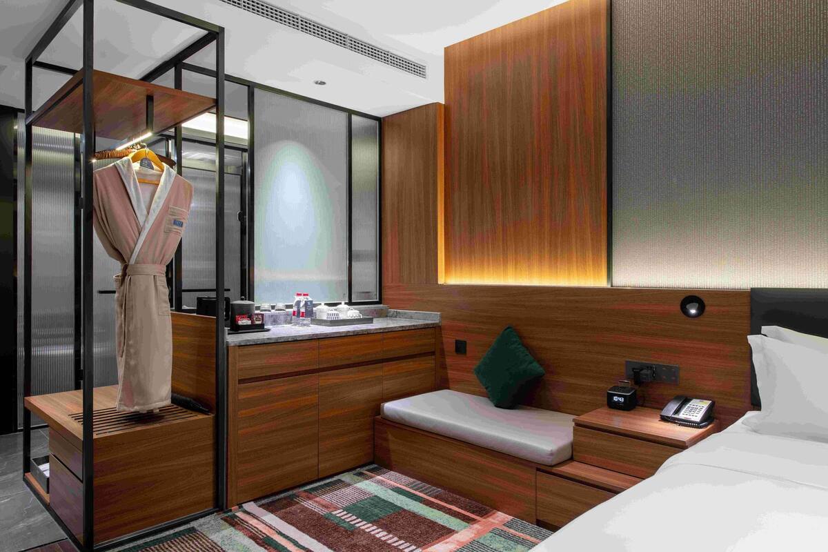 A compact hotel room with a wooden vanity, a small seating area, a bed with a pillow, a bedside table with a phone, and a mirror.