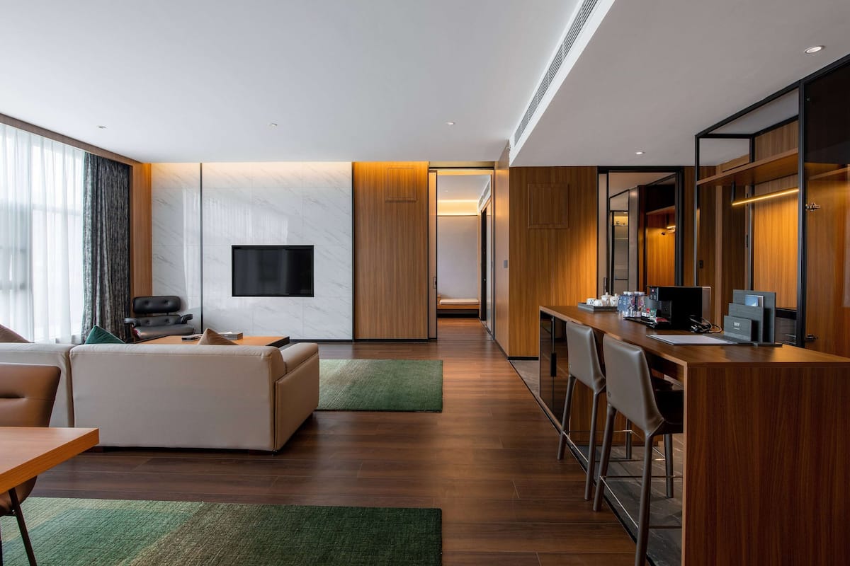 A modern hotel room with a sofa, a TV, a desk with chairs, and a wooden floor.