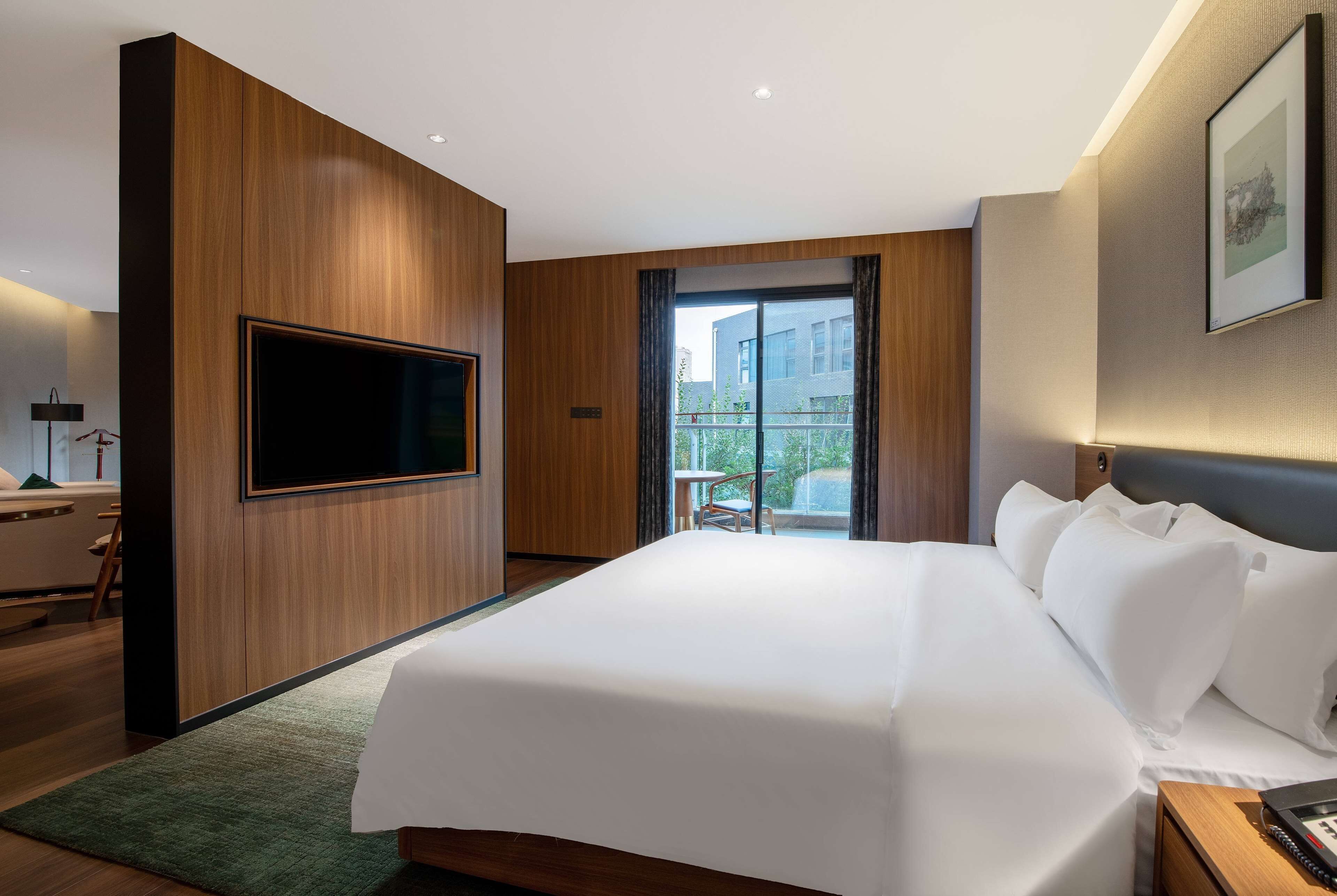 A hotel room with a large bed, a flat-screen TV mounted on the wall, a small table with a chair, and a window with a view of buildings outside.