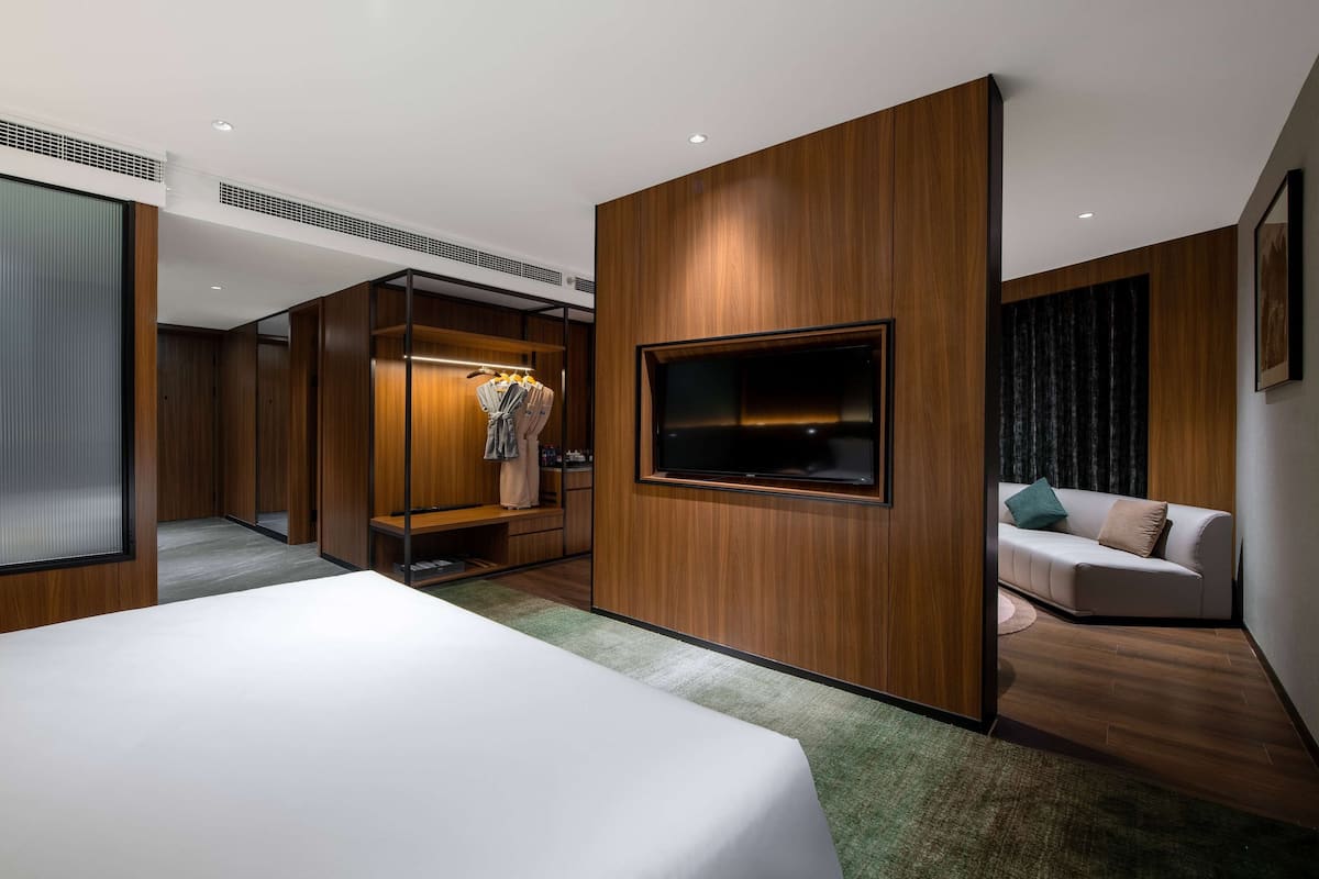 A modern hotel room with a large bed, a TV mounted on a wooden panel, a seating area with a sofa, and a walk-in closet.