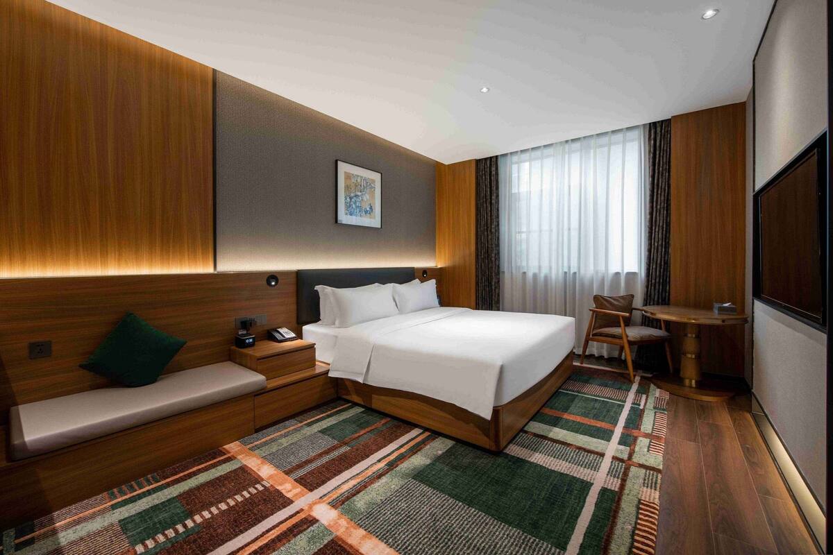 A hotel room with a large bed, a bedside table, a chair, a small desk, and a window with curtains.