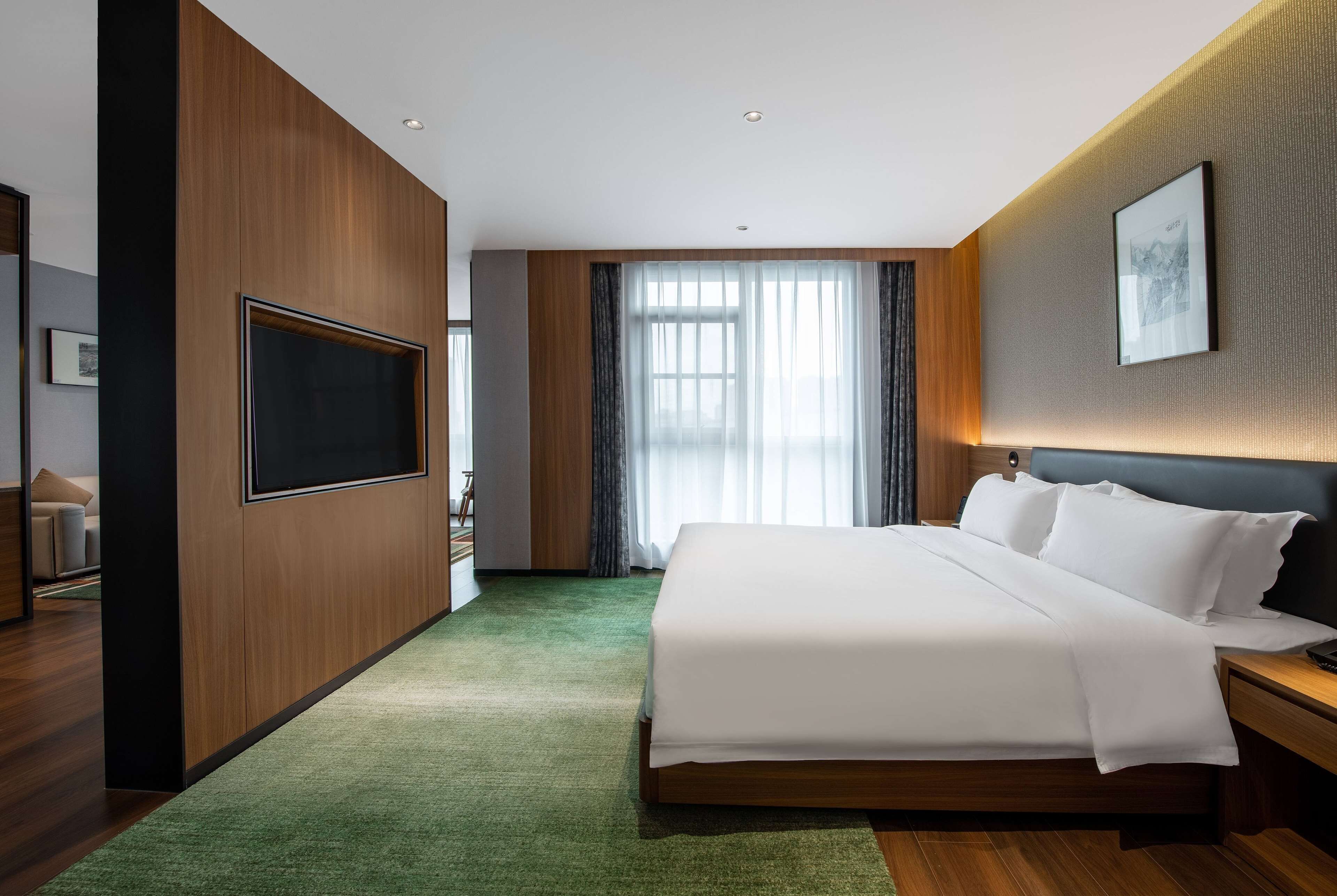 Una habitación de hotel moderna con una cama grande, un televisor de pantalla plana, una pared decorativa de madera y un ventanal con cortinas.
