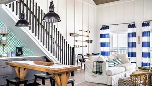 Een modern interieur met een houten eettafel, zwarte barkrukken, een witte bank met gestreepte kussens en een trap met zwarte leuningen.