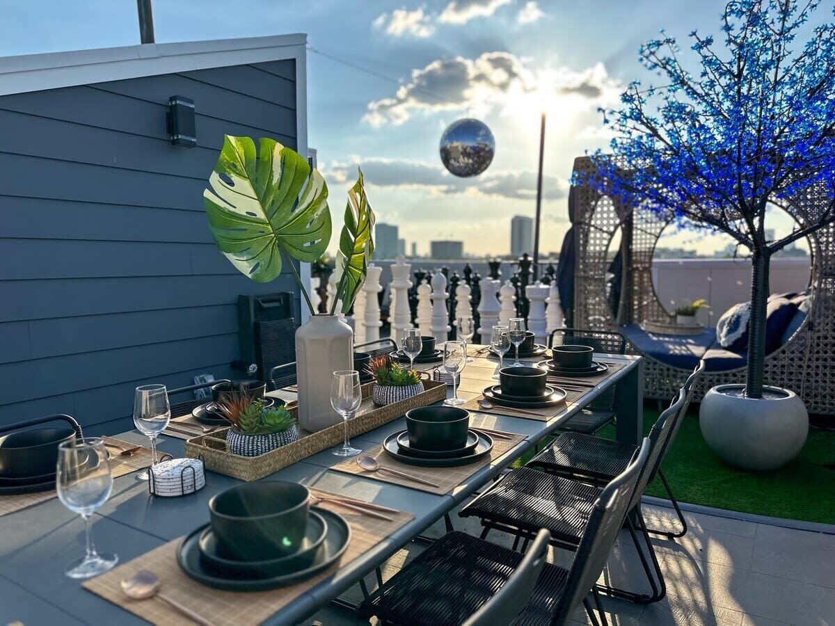 En tagterrasse med et langt bord dækket til et måltid, omgivet af stole, plantekasser og et dekorativt træ.