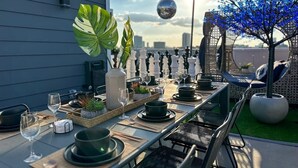 A rooftop dining area with a long table set for a meal, surrounded by chairs, potted plants, and a decorative tree. - The Oasis Checkmate HTX Rooftop (Houston)