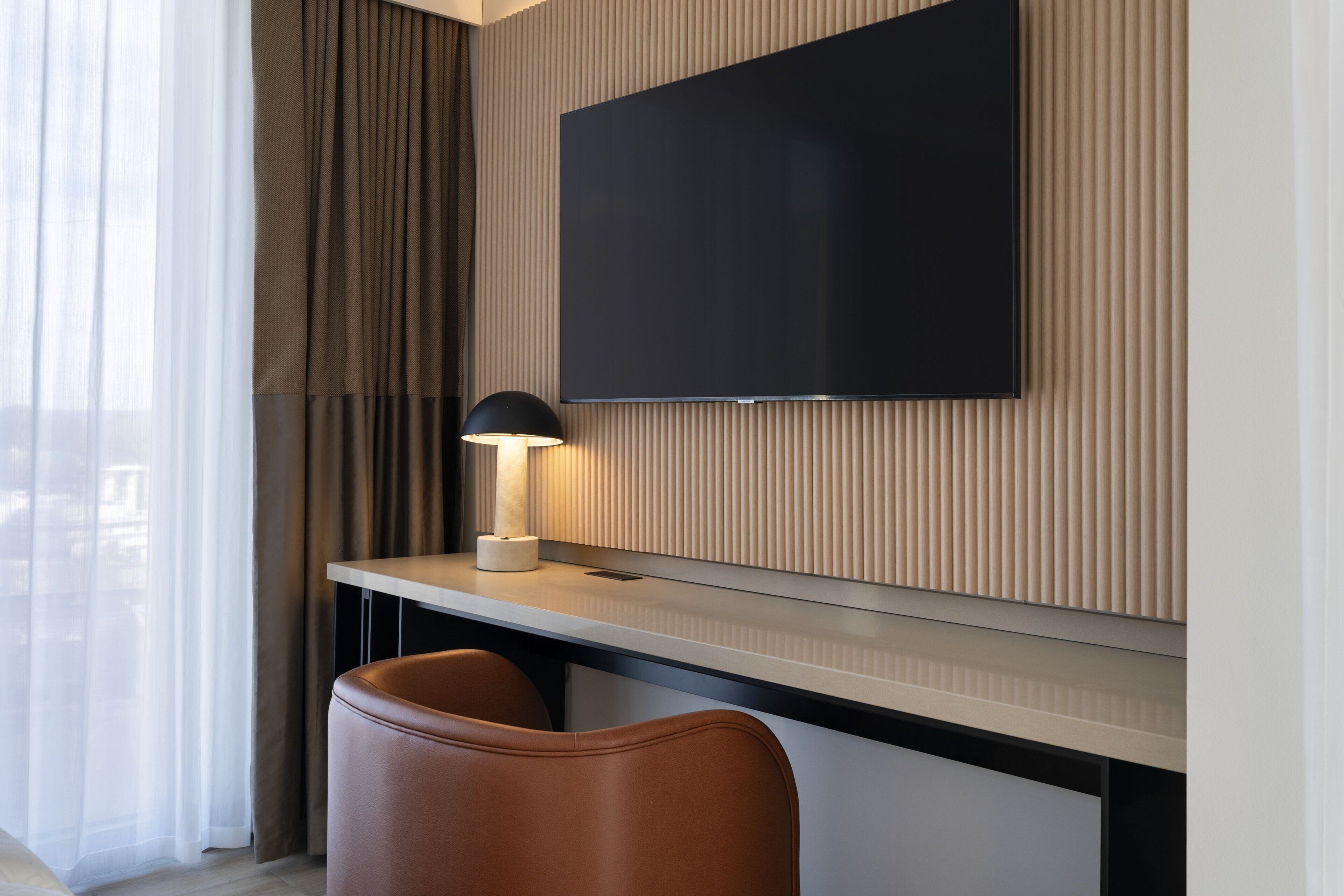Quarto de hotel moderno com televisão de tela plana fixada em uma parede de painel de madeira, uma escrivaninha com luminária e duas poltronas de couro.