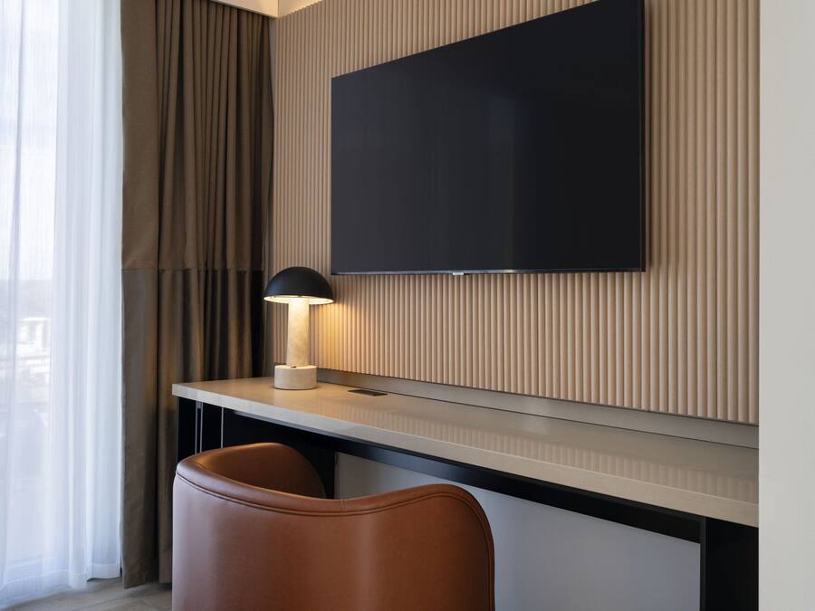 Quarto de hotel moderno com televisão de tela plana fixada em uma parede de painel de madeira, uma escrivaninha com luminária e duas poltronas de couro.